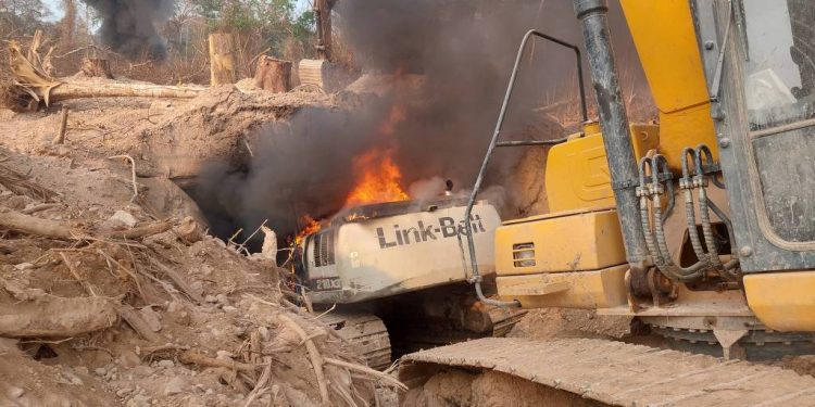 A imagem mostra uma escavadeira, da marca Link-Belt, pegando fogo em uma área de desmatamento. Há fumaça preta saindo da máquina e do solo ao redor, que está coberto de terra e troncos de árvores derrubadas. O ambiente é árido, com vegetação escassa ao fundo. Escavadeira usada pelos garimpeiros na Terra Indígena Sararé, no Mato Grosso, é destruída em operação do Ibama e da PRF