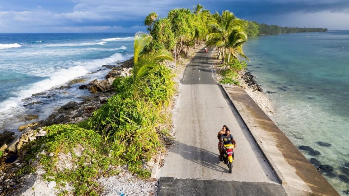 Um casal em uma motocicleta passa pelo ponto mais estreito da ilha de Fongafale, no atol de Funafuti, parte da nação insular Tuvalu. O Oceano Pacífico está à esquerda, e uma lagoa está à direita. A nação foi identificada como uma das mais vulneráveis do mundo às mudanças climáticas.