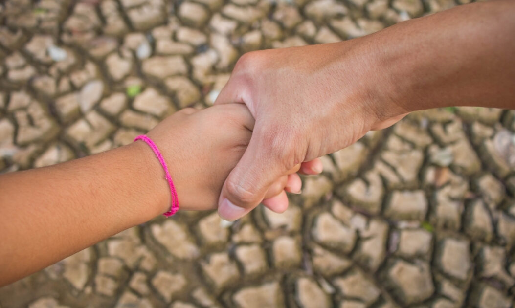 Mão adulta segurando a mão de uma criança sobre solo seco, simbolizando a vulnerabilidade diante das mudanças climáticas e o aumento de 22% no risco de mortalidade infantil. Foto: Canva.