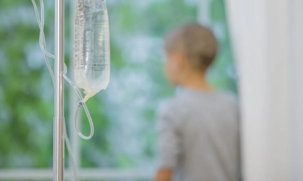 Frasco de soro hospitalar em primeiro plano com criança em tratamento desfocada ao fundo, simbolizando desafios do câncer infantil raro.
