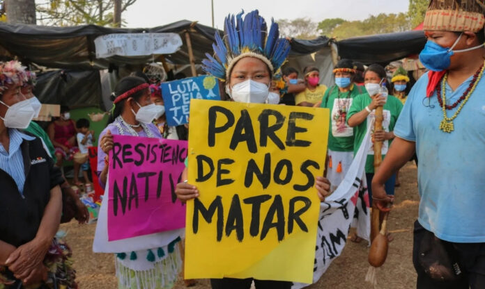 Indígenas em protesto exibem cartaz com os dizeres “pare de nos matar”, exigindo fim da violência contra ativistas ambientais.