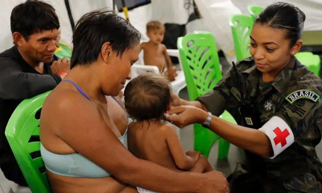 Criança Yanomami sendo atendida por profissional do Exército Brasileiro, com pais ao lado e sorriso mútuo durante ação de saúde indígena.