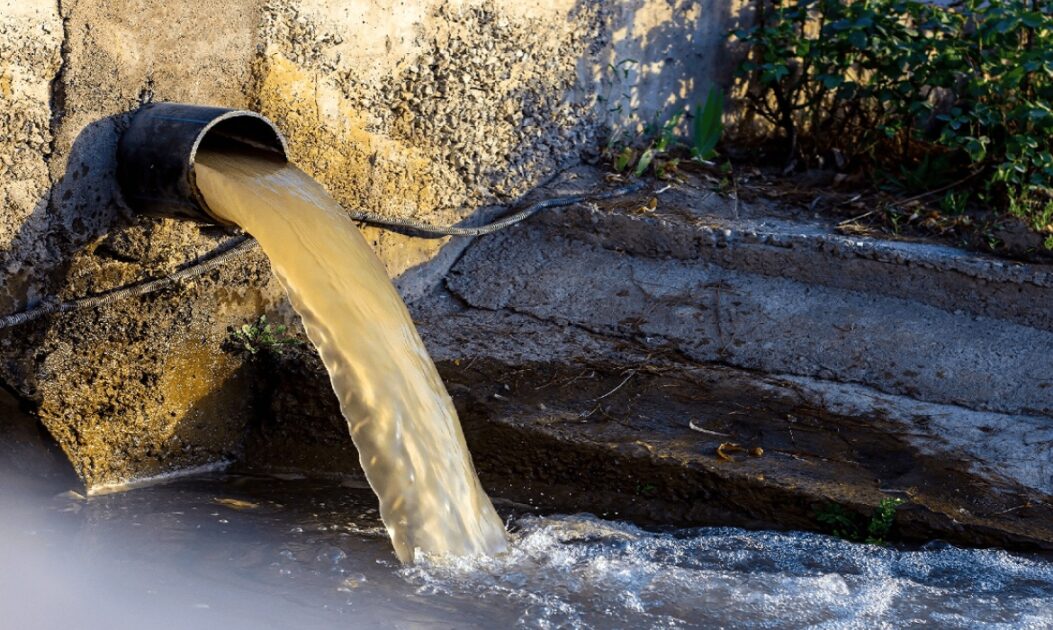 Água contaminada sendo despejada por cano em ambiente natural, ilustrando poluição da água por PFAS.