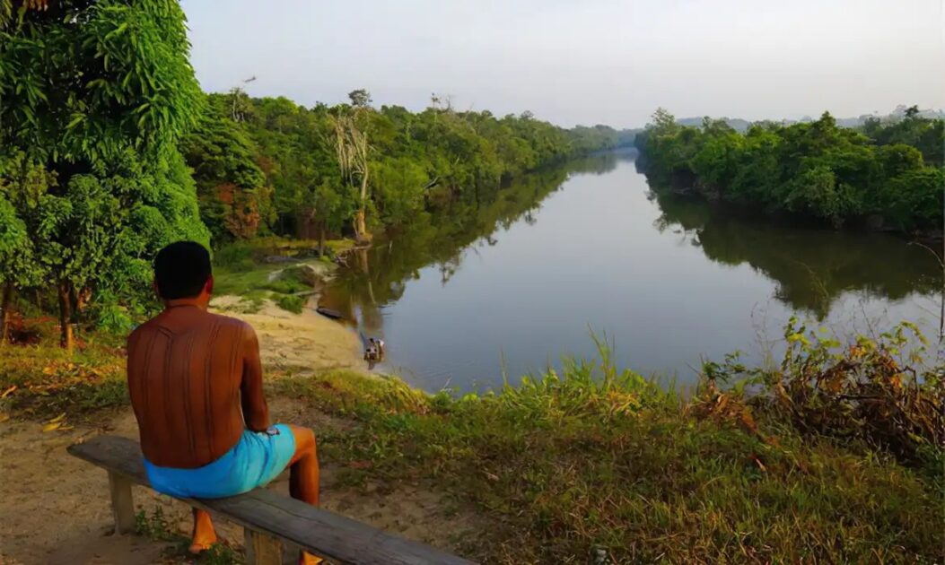 Indígena sentado diante de um lago na floresta; o marco temporal coloca territórios em risco.
