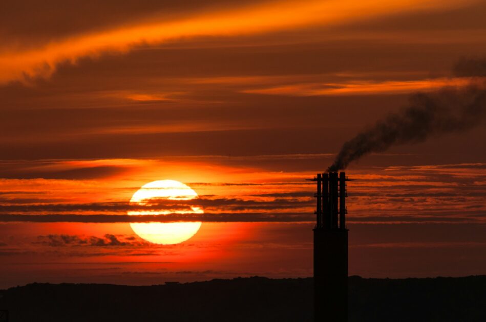 Chaminé industrial liberando fumaça sob o sol poente, ilustrando os danos ambientais provocados por emissões de gases poluentes. 