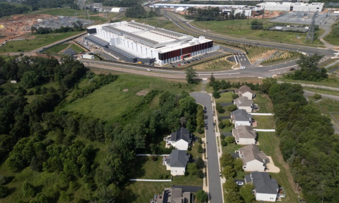 Imagem aérea de Briarfield Estates e Hiddenwood in Northern Virginia que mostra a imensidão e repetição dos galpões de data centers com telhados metálicos, linhas de transmissão elétrica e torres de resfriamento, contrastando com bairros ou áreas verdes ao redor.