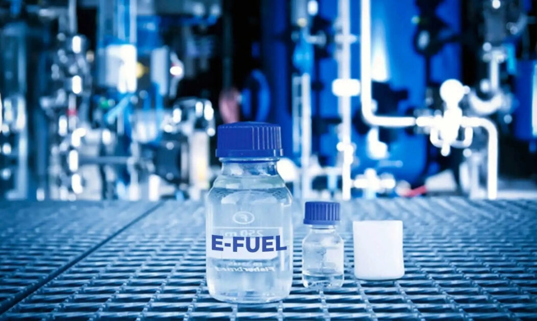 Frasco de e-fuel em primeiro plano com fábrica ao fundo representa a produção de combustíveis sintéticos e o avanço dos combustíveis do futuro.