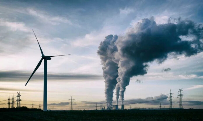 Turbina eólica em primeiro plano com chaminé industrial ao fundo, representando a geopolítica da energia e a transição energética entre fontes fósseis e renováveis.